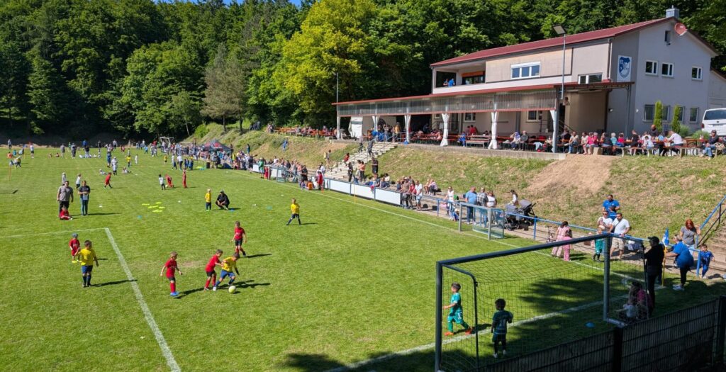 Kinder spielen ein Fußballspiel auf einer Wiese, während die Zuschauer an einem sonnigen Tag vom Spielfeldrand und einem nahe gelegenen, von Bäumen umgebenen Clubhaus aus zusehen.